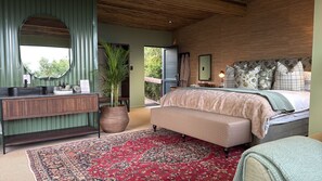 A bedroom with a large bed, a bedside table, a mirror, a potted plant, and a view of the outdoors.