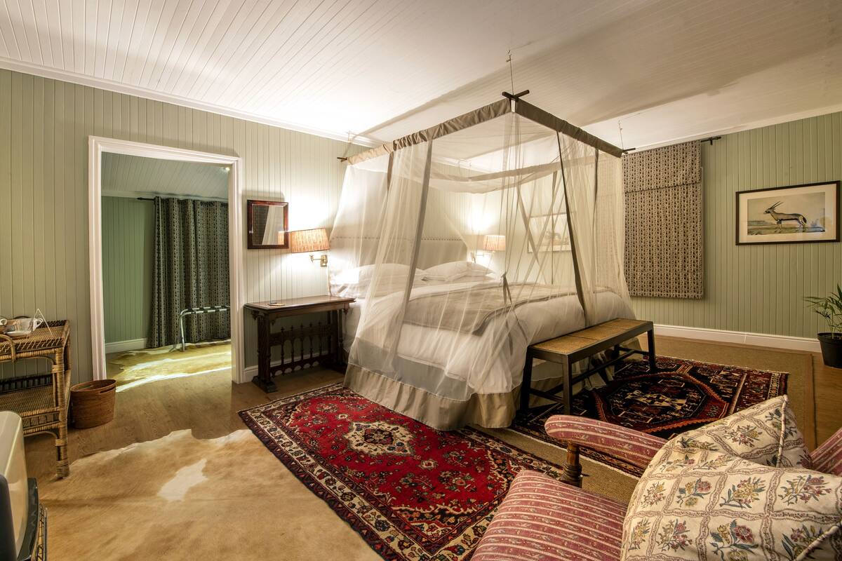 A traditional bedroom with a canopy bed, a sitting area with a red patterned rug, and a view of the hallway.