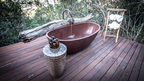 A wooden deck with a large, round, brown bathtub, a wooden stool with bottles, and a rack with towels.