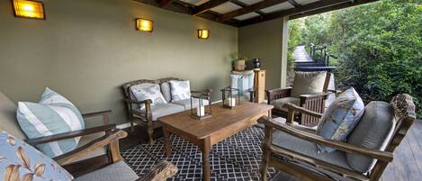 A covered patio with wooden furniture, including a sofa and chairs, a wooden table, and two wall-mounted lamps.