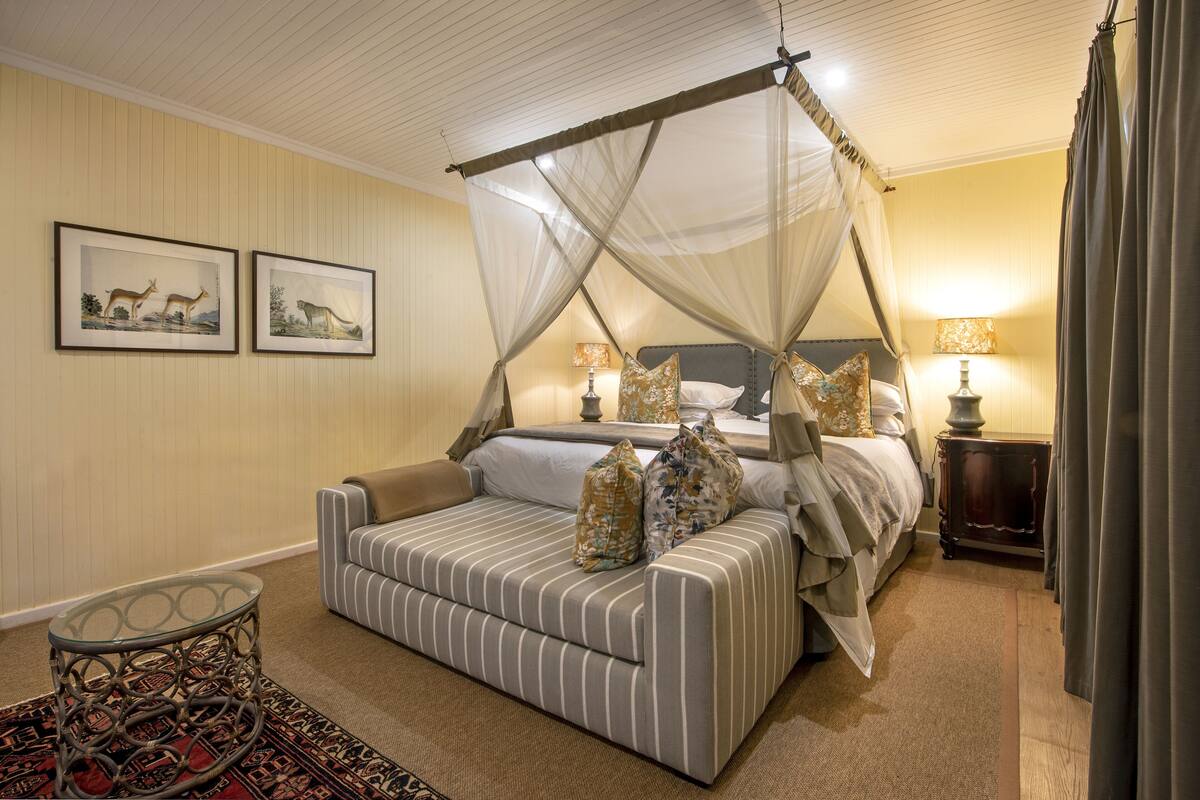 A four-poster canopy bed with a grey and white striped sofa, two framed animal paintings, and a small round table with a patterned rug.