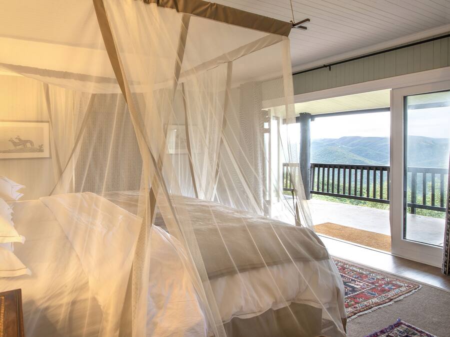 A four-poster bed with sheer curtains, a bedside lamp, and a view of a mountainous landscape through a balcony.