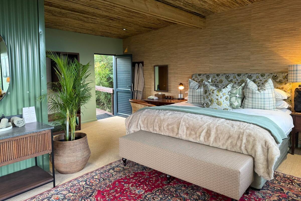 A bedroom with a bed, a bench, a nightstand, a mirror, a potted plant, and a view of the outdoors.
