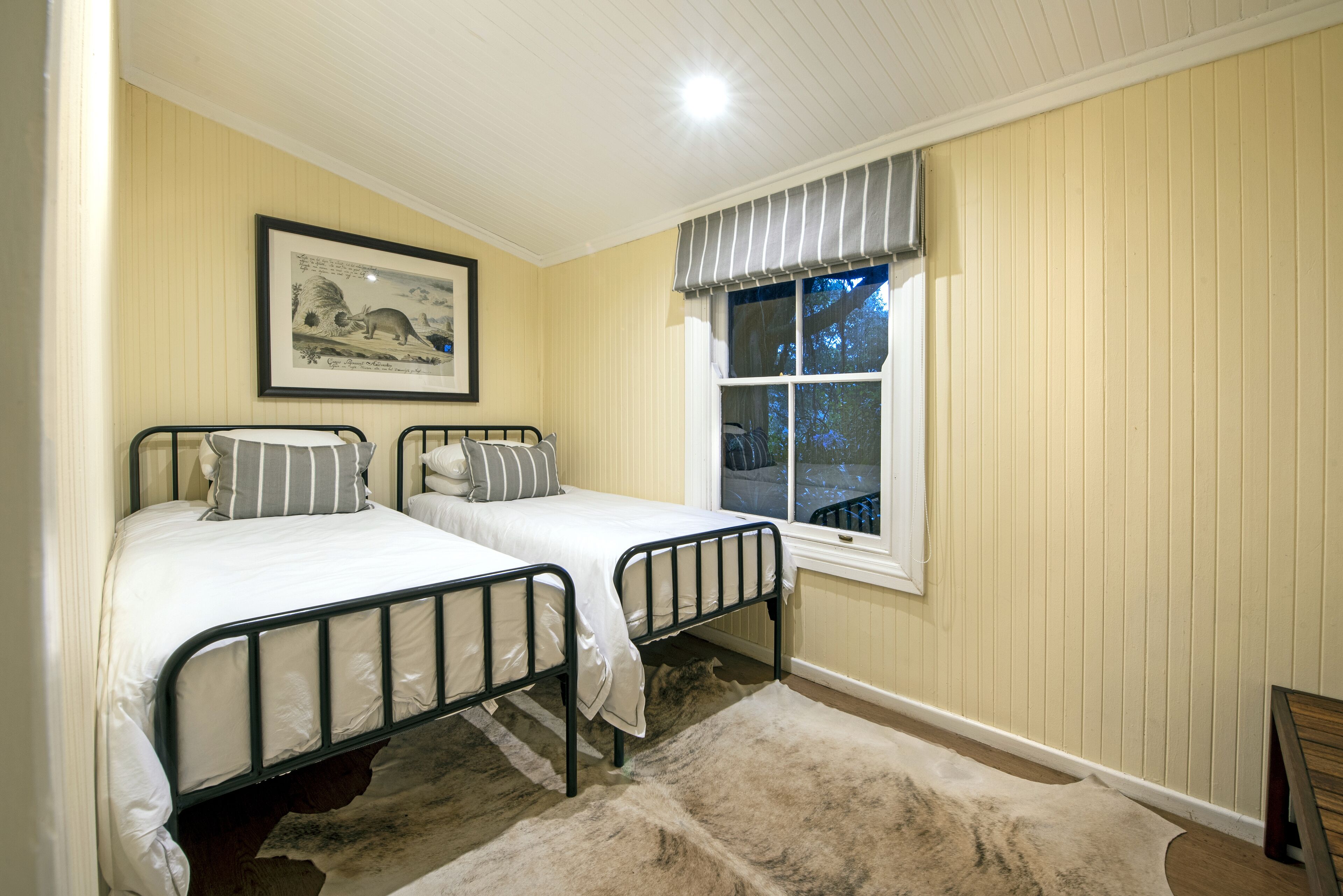 A bedroom with two single beds, a framed picture on the wall, a window with a view, and a rug on the floor.
