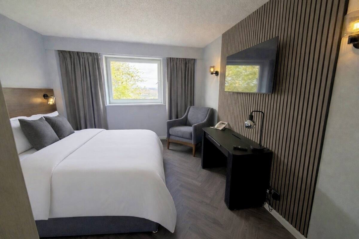 A hotel room with a bed, a desk, a chair, a TV mounted on the wall, and a window with curtains.
