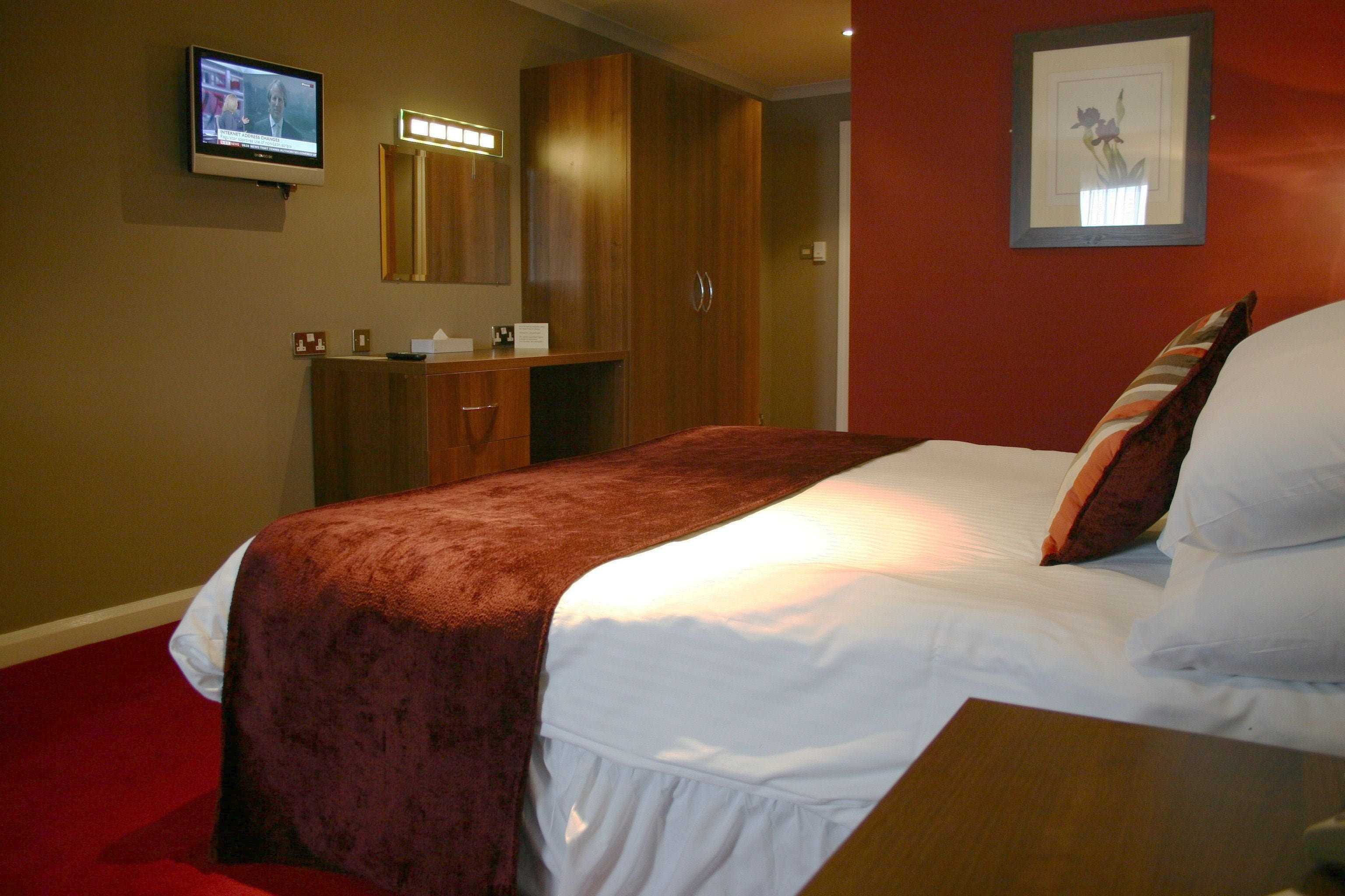 Una habitación de hotel con una cama, un televisor montado en la pared, un armario de madera y un cuadro enmarcado.