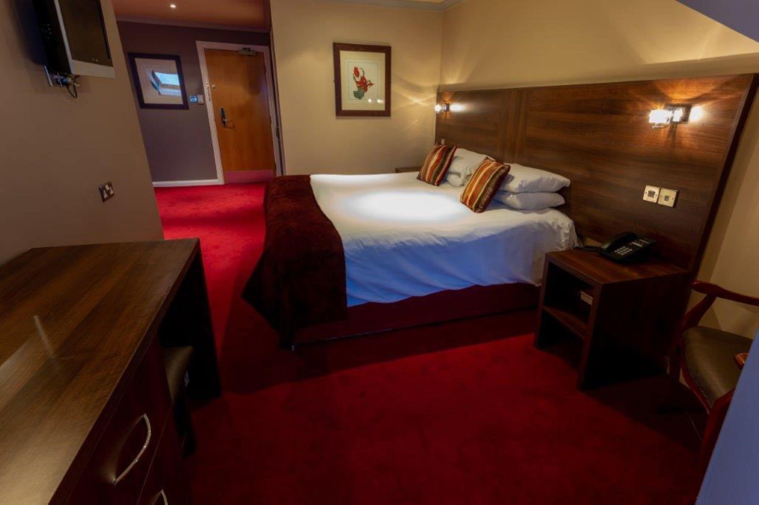 A hotel room with a large bed, a wooden headboard, a desk with a chair, and a red carpet.