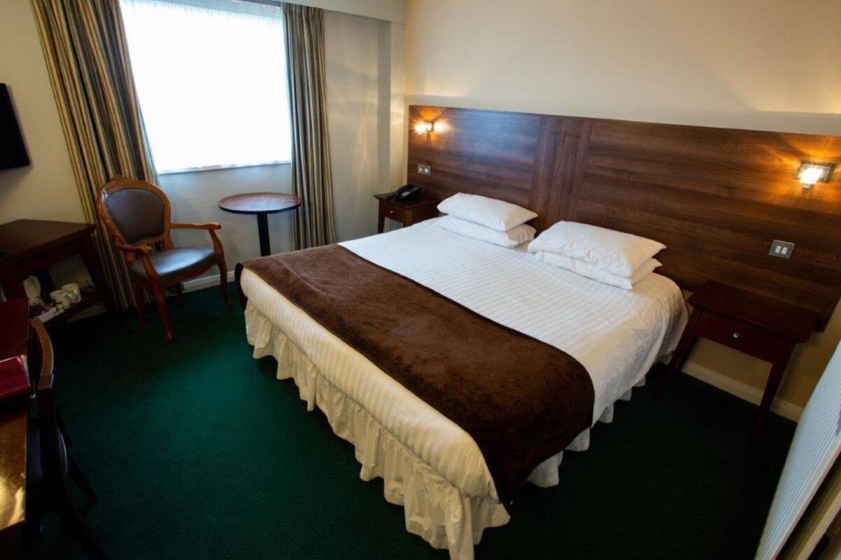 A hotel room with a large bed, a bedside table, a chair, a small table, and a window with curtains.