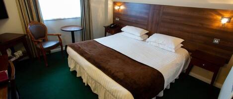 A hotel room with a large bed, a bedside table, a chair, a small table, and a window with curtains.