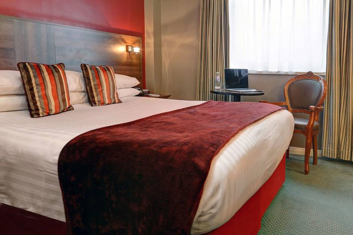 A hotel room with a large bed, striped pillows, a red throw blanket, a wooden headboard, a small table with a laptop, a chair, and a window with curtains.