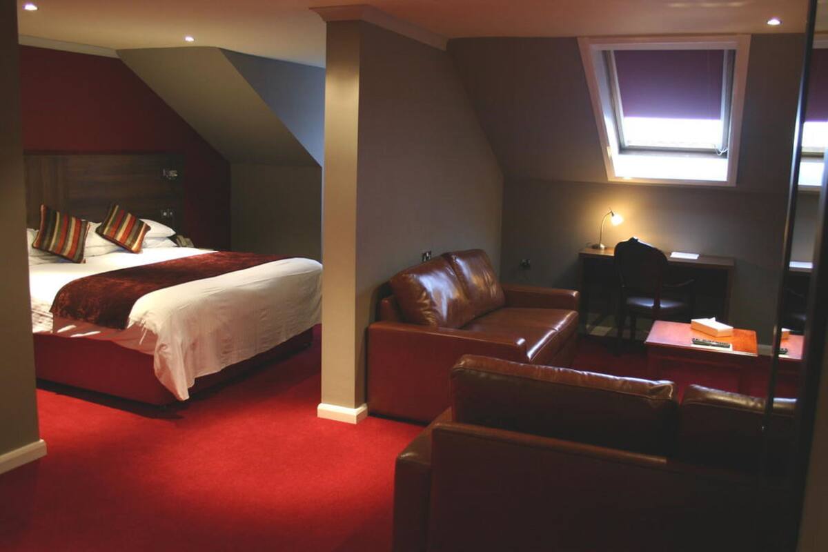 A bedroom with a bed, a leather sofa, a desk with a lamp, and a window.