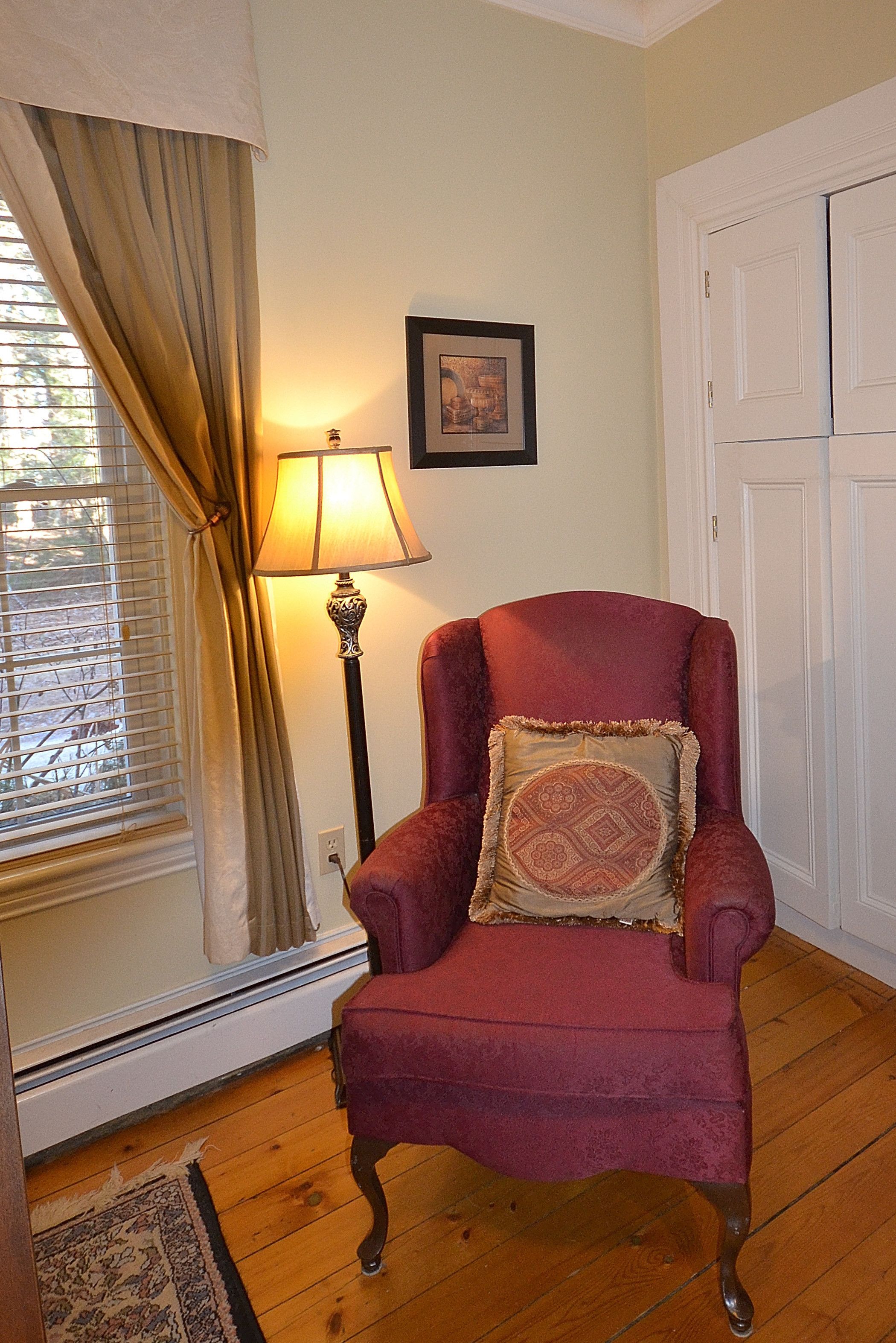 A room with a red armchair, a floor lamp, a window with curtains, and a door.