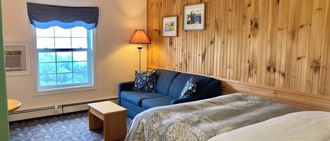 A room with a bed, a sofa, a small table, a lamp, and a window with a view of trees.