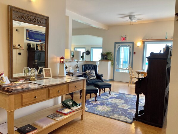 Une pièce bien éclairée, dotée d’un grand miroir, d’un bureau en bois avec des livres, d’un fauteuil bleu et d’un piano.