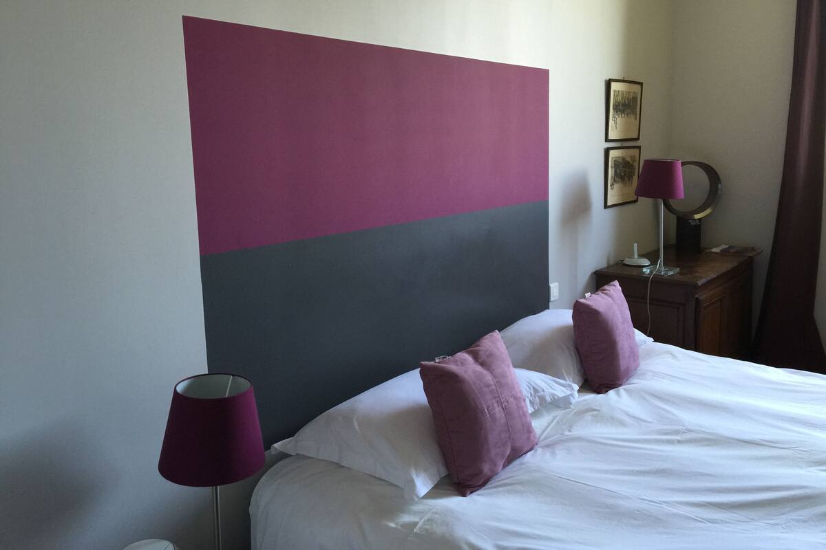 Ein Hotelzimmer mit einem großen Bett, zwei Kissen, einem Nachttisch mit Lampe und einer Wand mit einem großen abstrakten Gemälde.