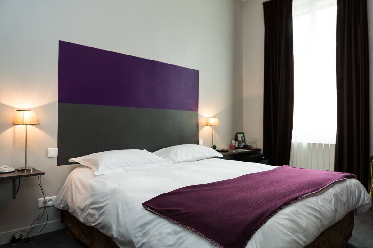 Ein Hotelzimmer mit einem großen Bett, zwei Nachttischlampen, einem Nachttisch und einem Fenster mit Vorhängen.