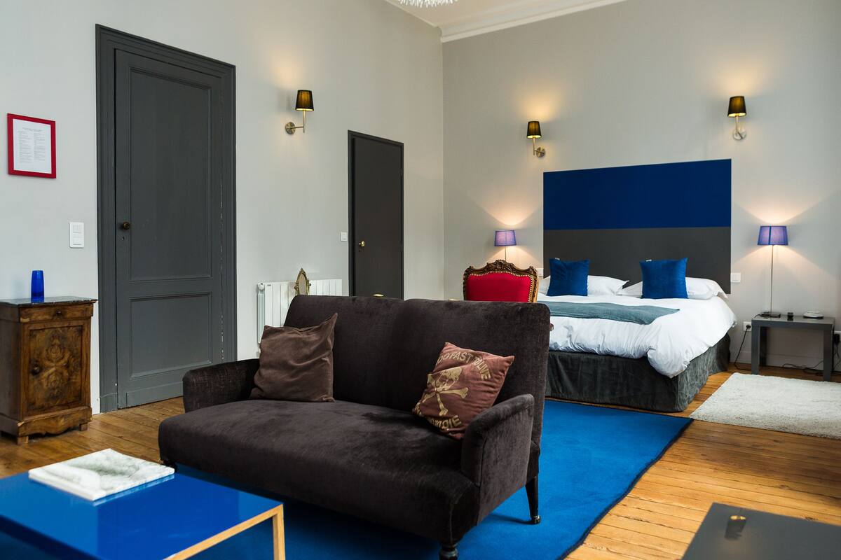 Ein modernes Hotelzimmer mit einem dunkelbraunen Sofa, einem Bett mit blau-weißer Bettwäsche und zwei Nachttischlampen.