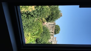 Vista a través de una ventana de un edificio de fachada de piedra y ventanas de arco, rodeado de abundante vegetación.
