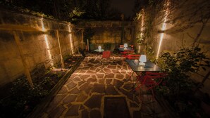 Un patio de piedra con mesas y sillas, rodeado de plantas y muros.