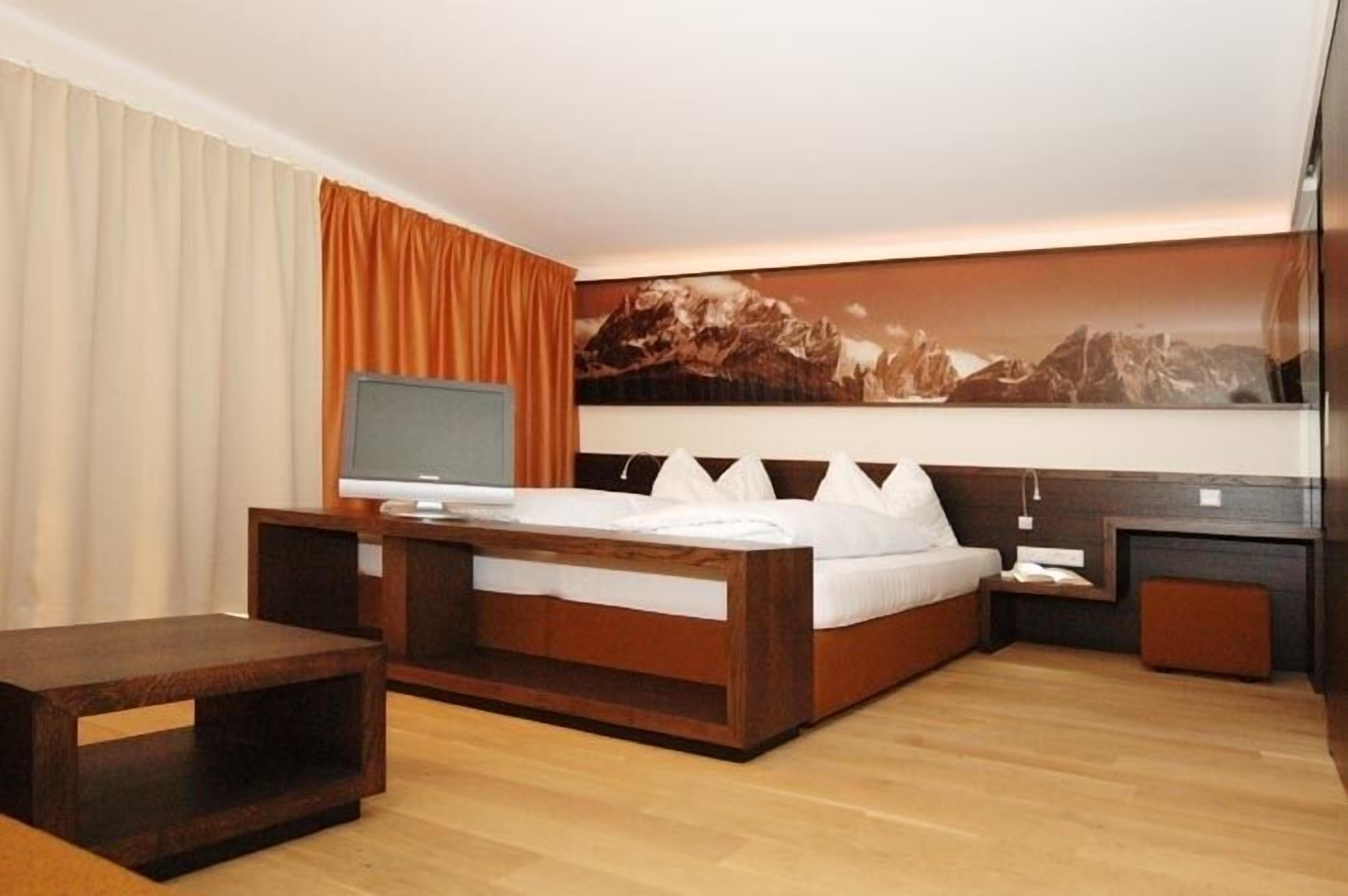 Una habitación de hotel moderna con una cama de madera, un televisor de pantalla plana y un cuadro grande de un paisaje montañoso en la pared.