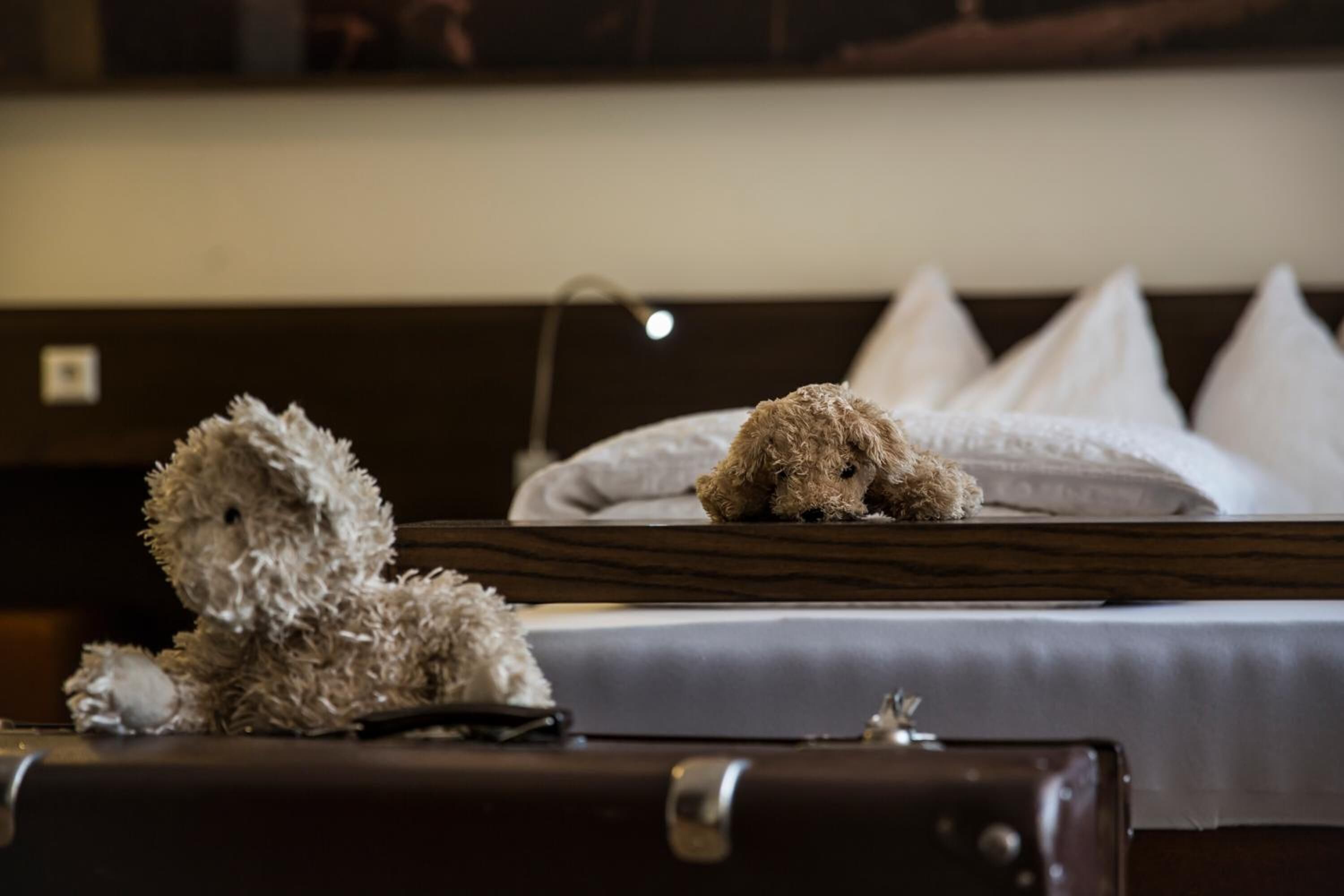 A hotel room with a bed, pillows, a teddy bear, and a suitcase.
