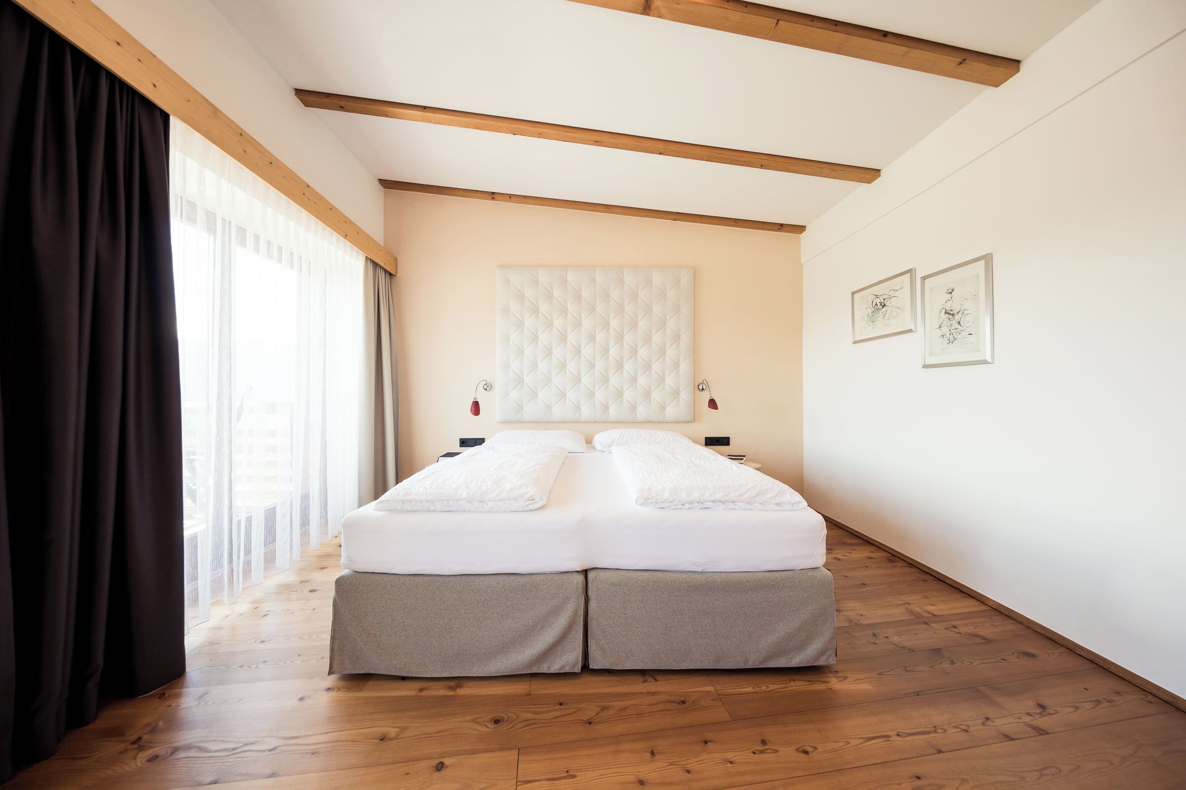 A bedroom with a bed, wooden beams, a window with curtains, and artwork on the wall.