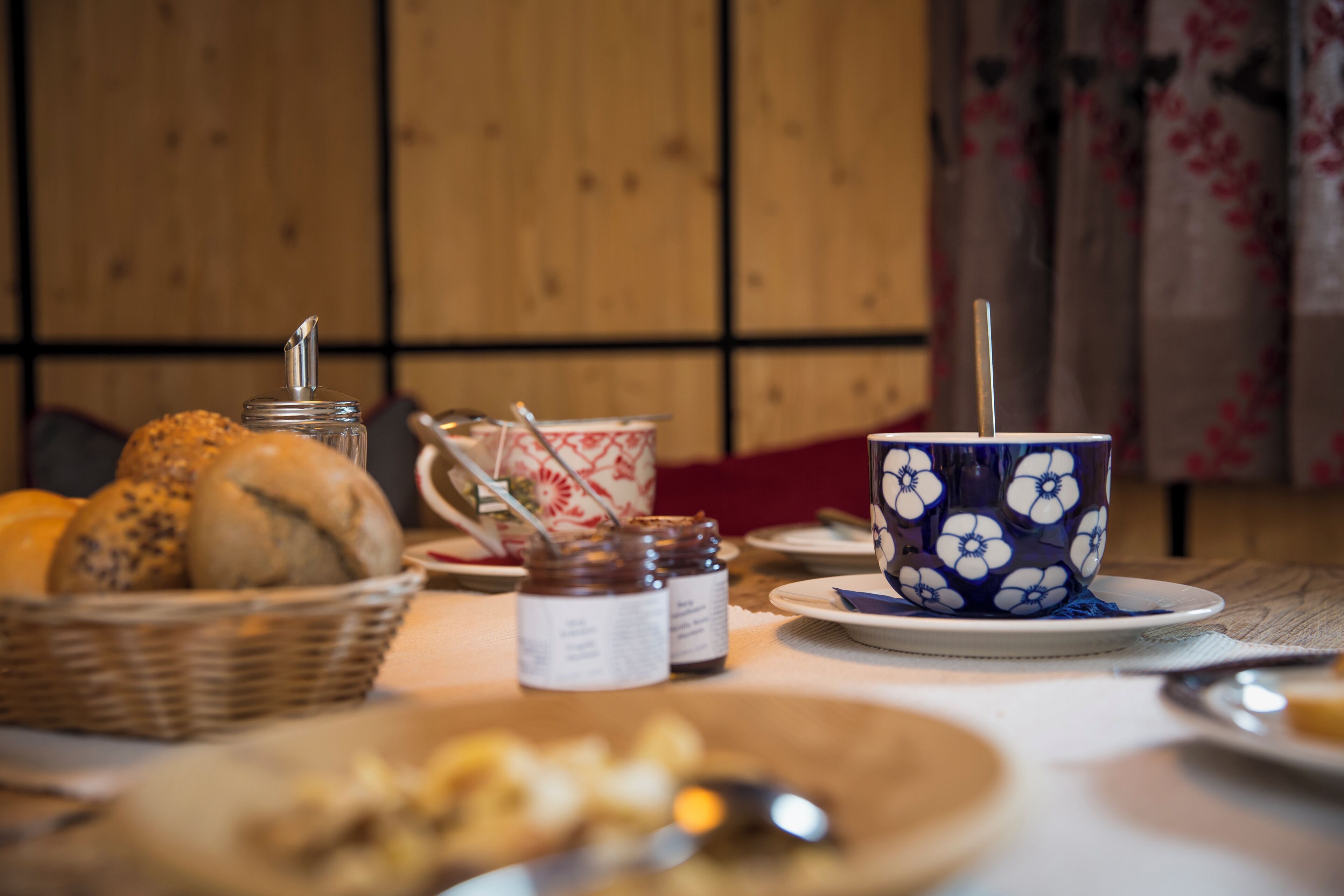 Ein Tisch mit einem Brotkorb, einer Tasse mit floralem Muster und einem Marmeladenglas.