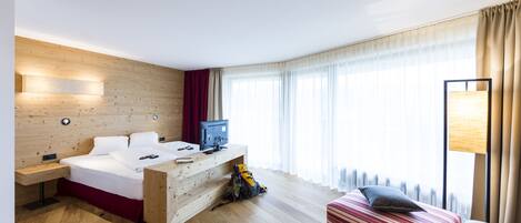A modern hotel room with a wooden headboard bed, a desk with a computer, a striped sofa, and a large window with curtains.