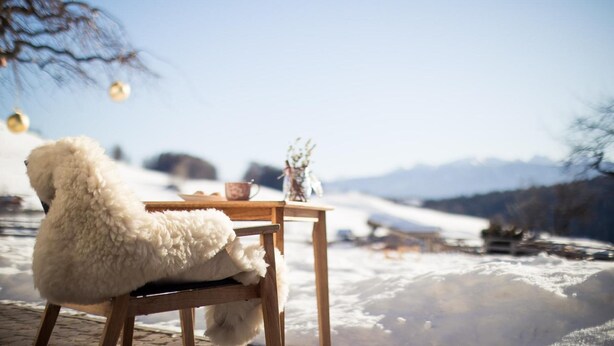 A wooden table with a cup and a vase, a fluffy white chair, and a snowy landscape in the background. - Hotel Dolomitenblick (Terento)
