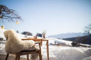 A wooden table with a cup and a vase, a fluffy white chair, and a snowy landscape in the background. - Hotel Dolomitenblick (Terento)