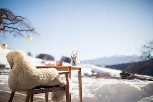 Una mesa de madera con una taza y un florero, una silla blanca y mullida, y un paisaje nevado al fondo.