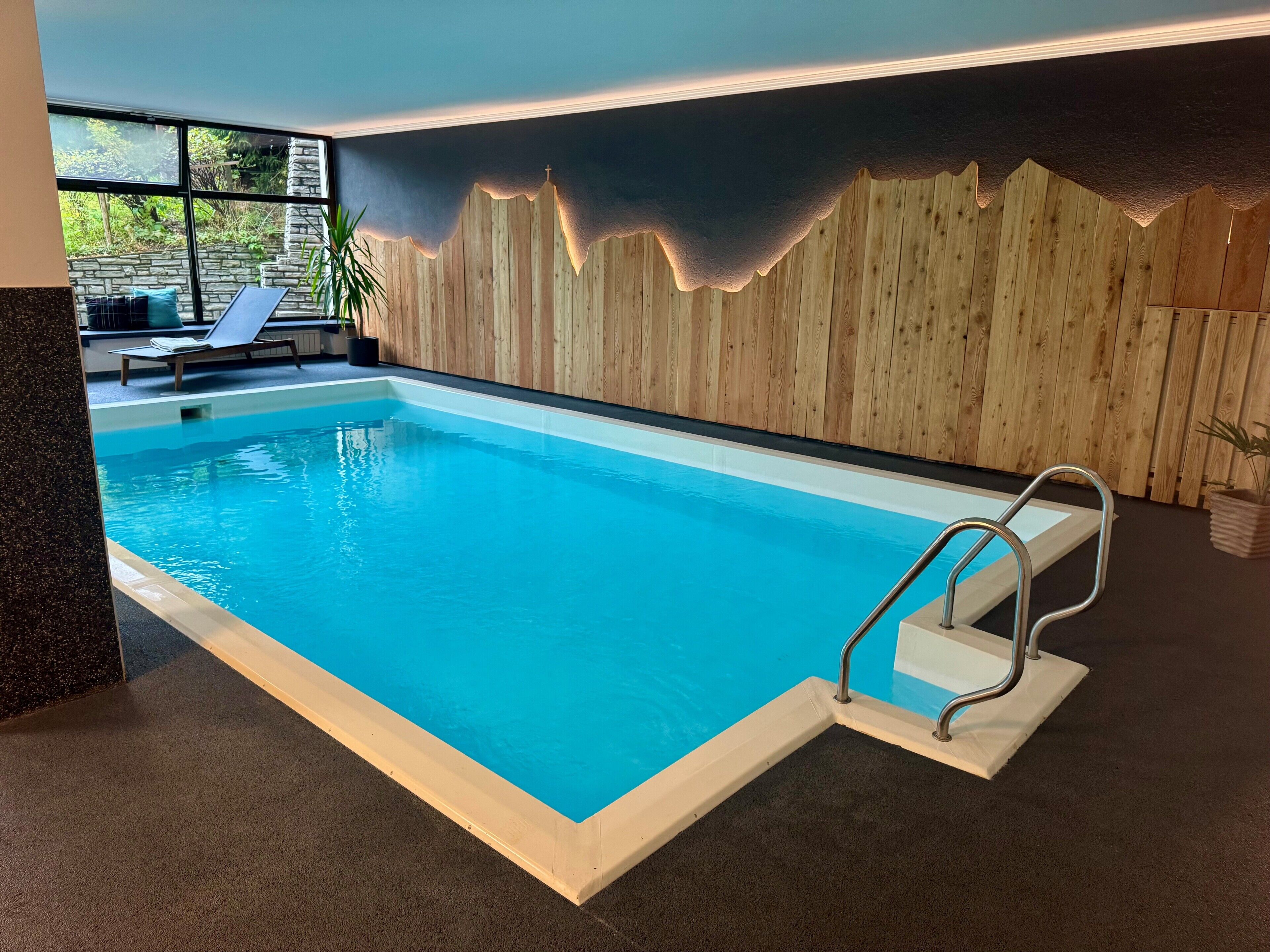 Una piscina interior con pared de madera, escalera metálica y zona de estar con vistas a zonas verdes.