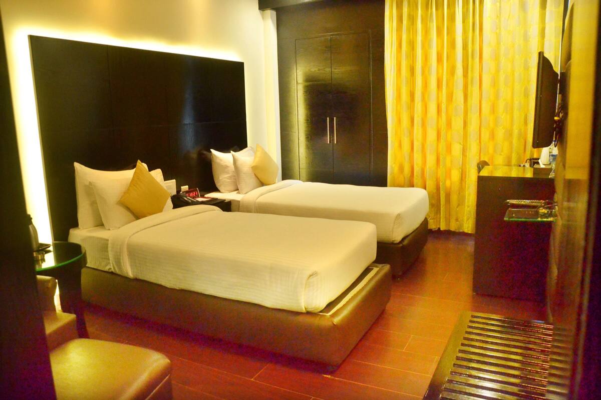 A hotel room with two beds, a large dark headboard, a wardrobe, and a bathroom with a glass shower enclosure.