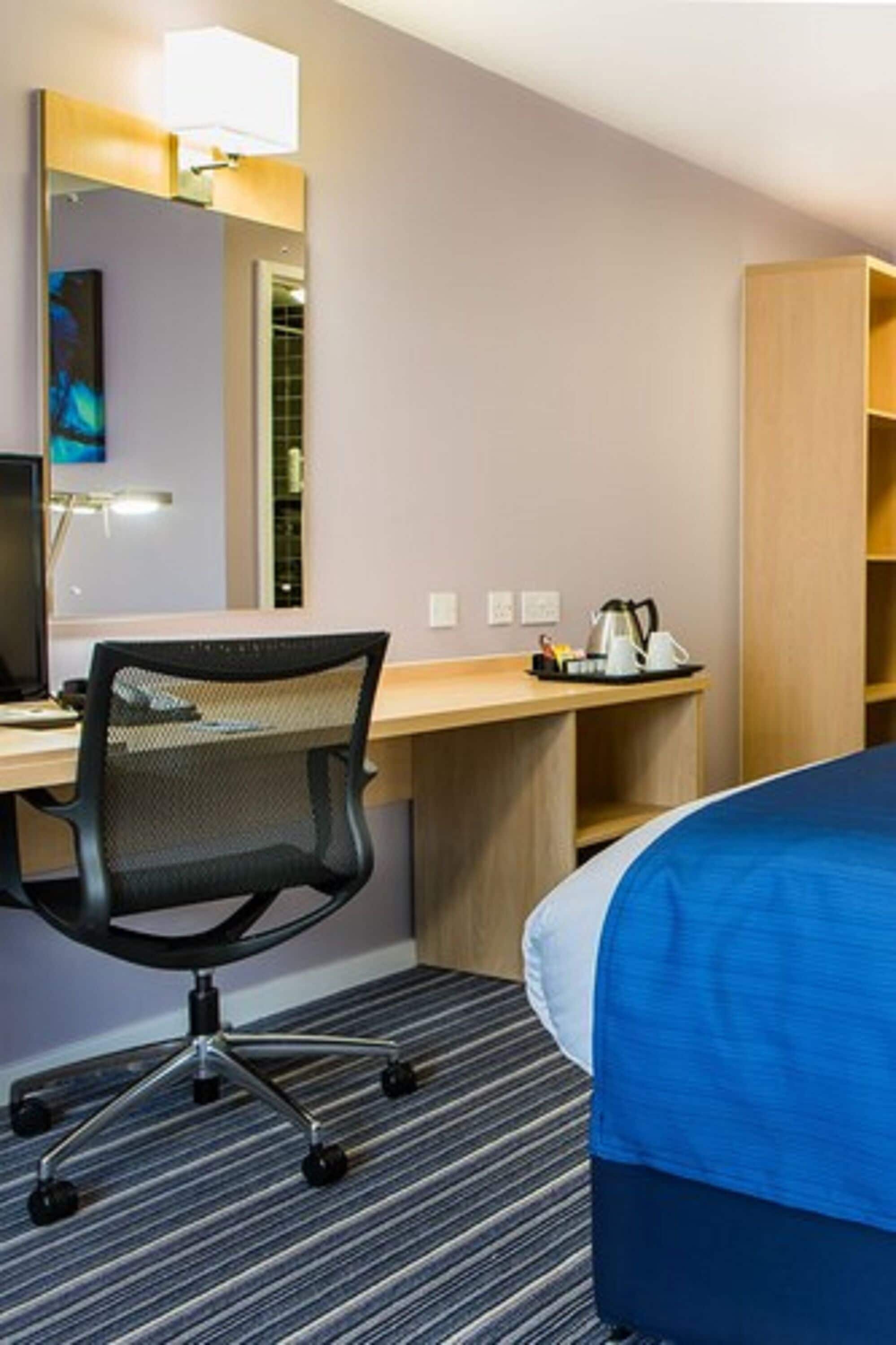 A hotel room with a bed, a desk with a computer and chair, a mirror, and a shelf with a kettle and other items.