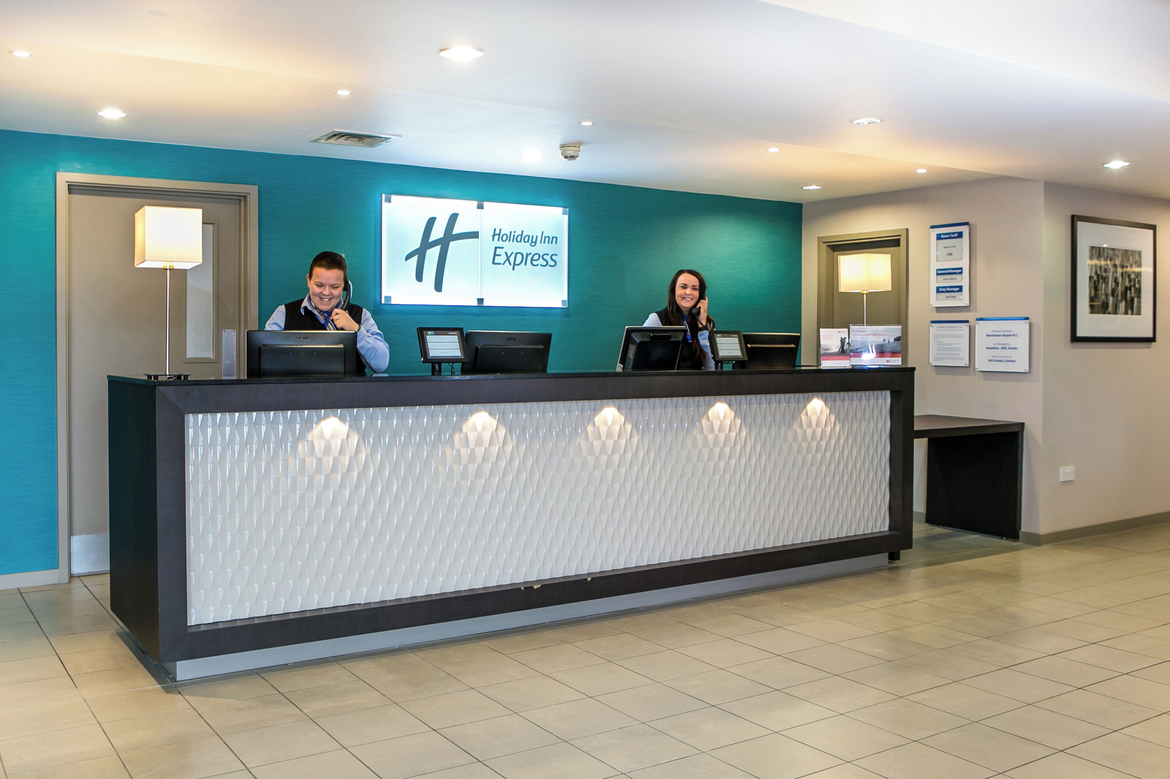Ein Empfangsschalter mit Holiday Inn Express Schild, zwei Rezeptionisten und einem modernen Interieur mit einer Türkis-farbenen Akzentwand.