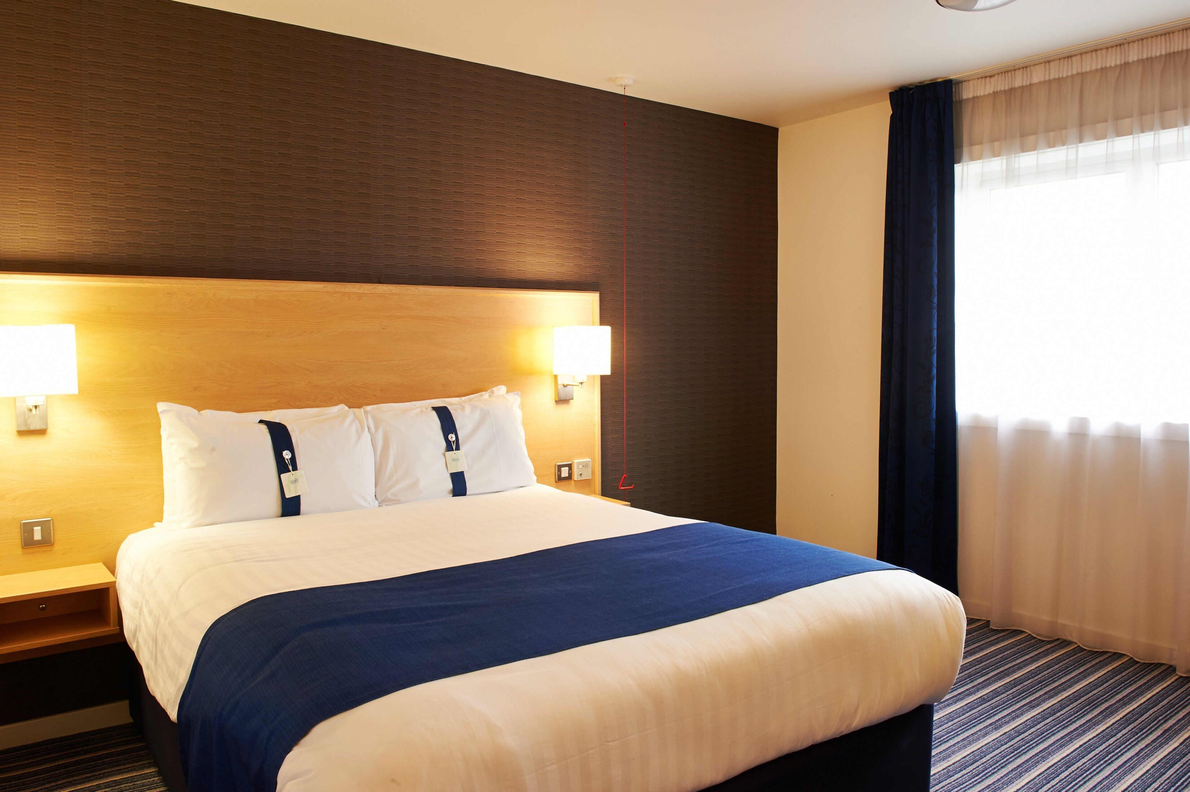 Ein Hotelzimmer mit einem großen Bett, zwei Nachttischlampen, einem hölzernen Kopfteil und einem Fenster mit Vorhängen.