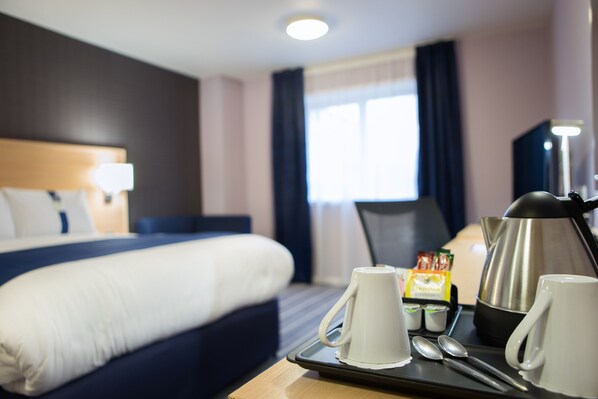 Une chambre d’hôtel avec un lit bien fait, un plateau avec des tasses et une bouilloire, et une fenêtre avec des rideaux.