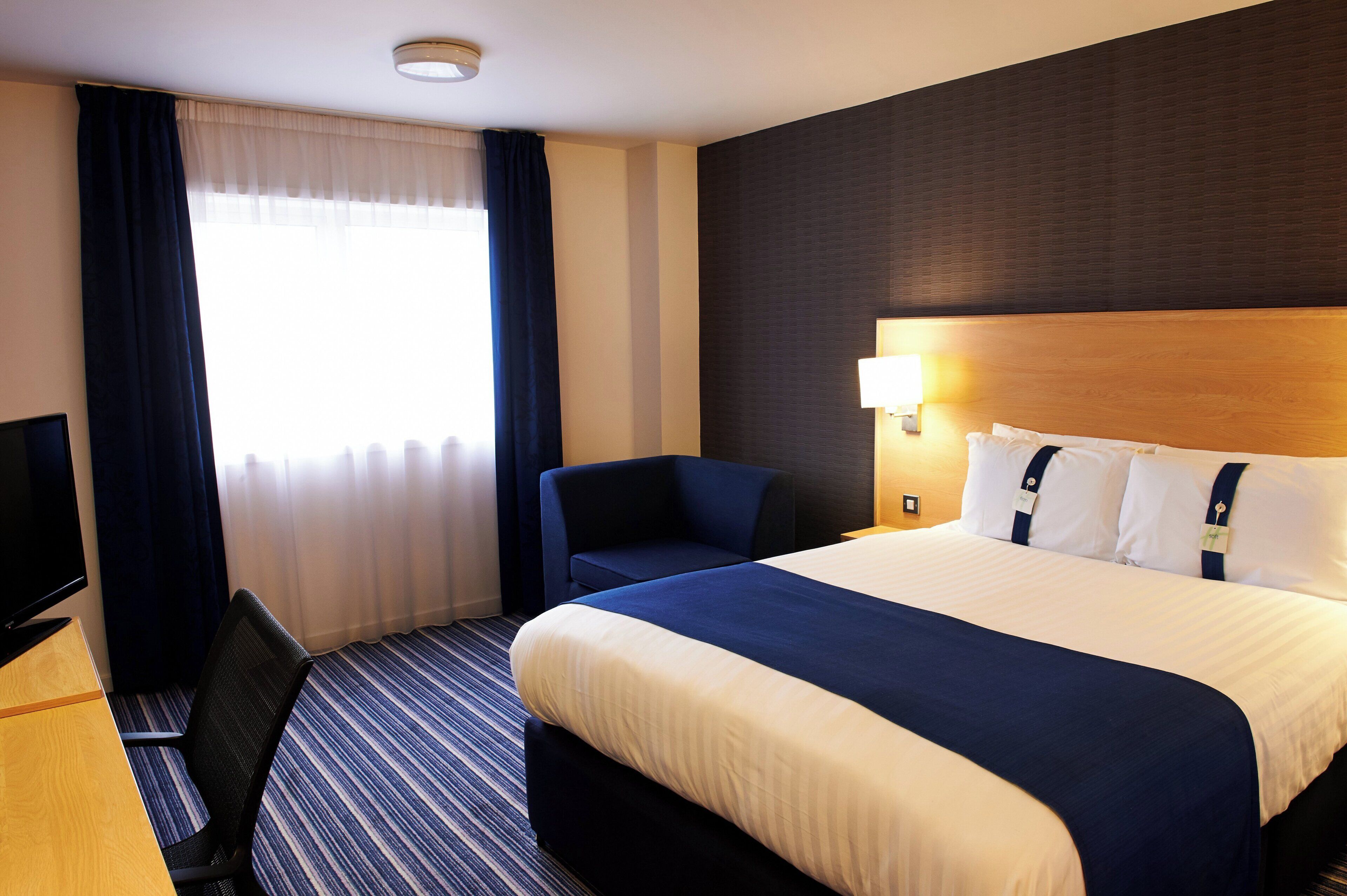 Una habitación de hotel con una cama grande, un escritorio con televisor, una silla y una ventana con cortinas.