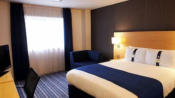 Quarto de hotel com uma cama grande, escrivaninha com televisão, cadeira e janela com cortinas.