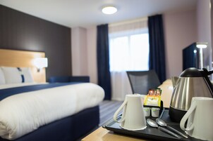 A hotel room with a neatly made bed, a tray with cups and a kettle, and a window with curtains.