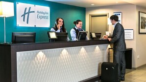 Eine Rezeption eines Holiday Inn Express mit drei Mitarbeitern, einem Gast mit Koffer und einer türkis-farbenen Wand mit dem Hotel-Logo.