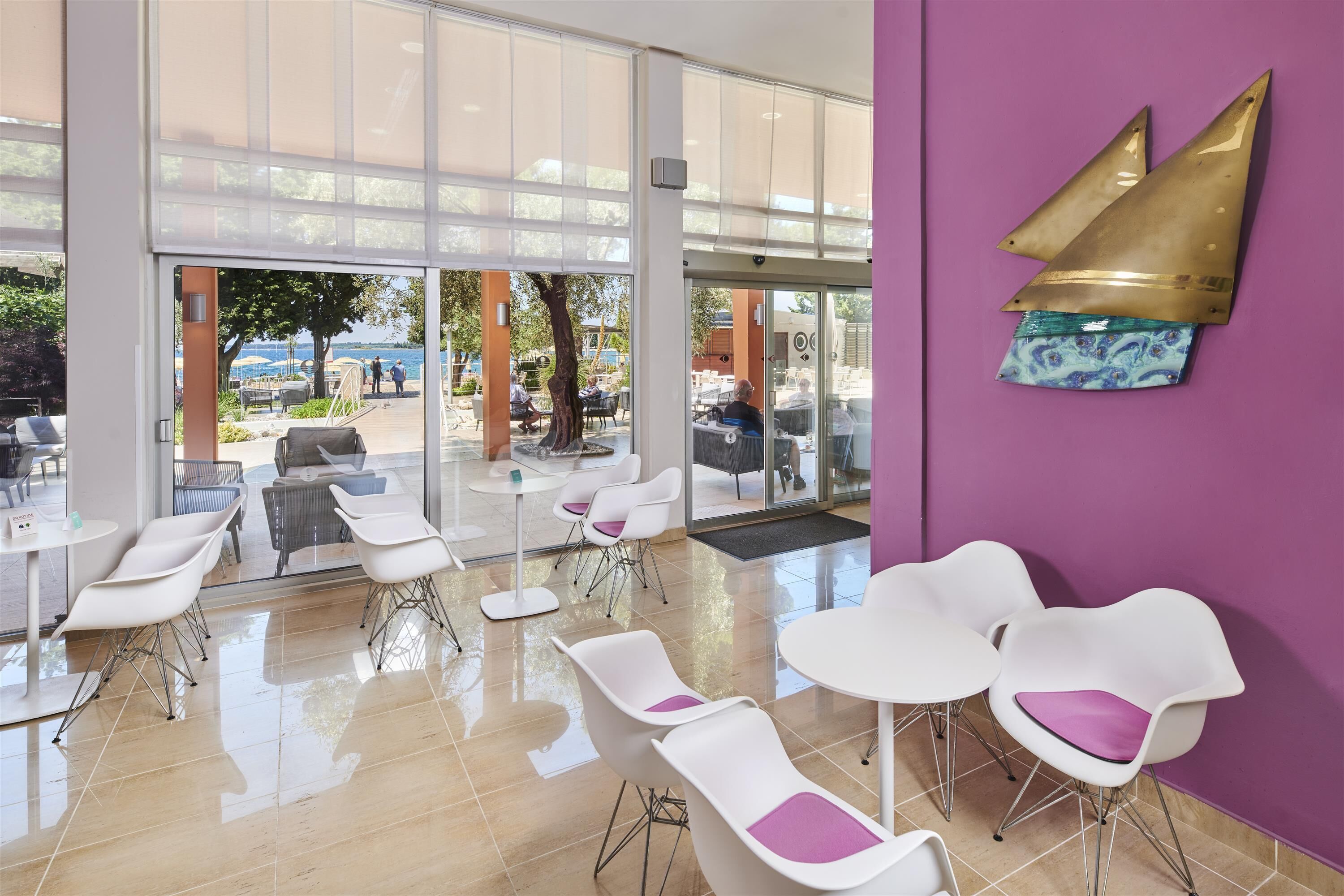 Un espacio interior moderno con sillas y mesas blancas, ventanas amplias y una pared morada con un adorno metálico.