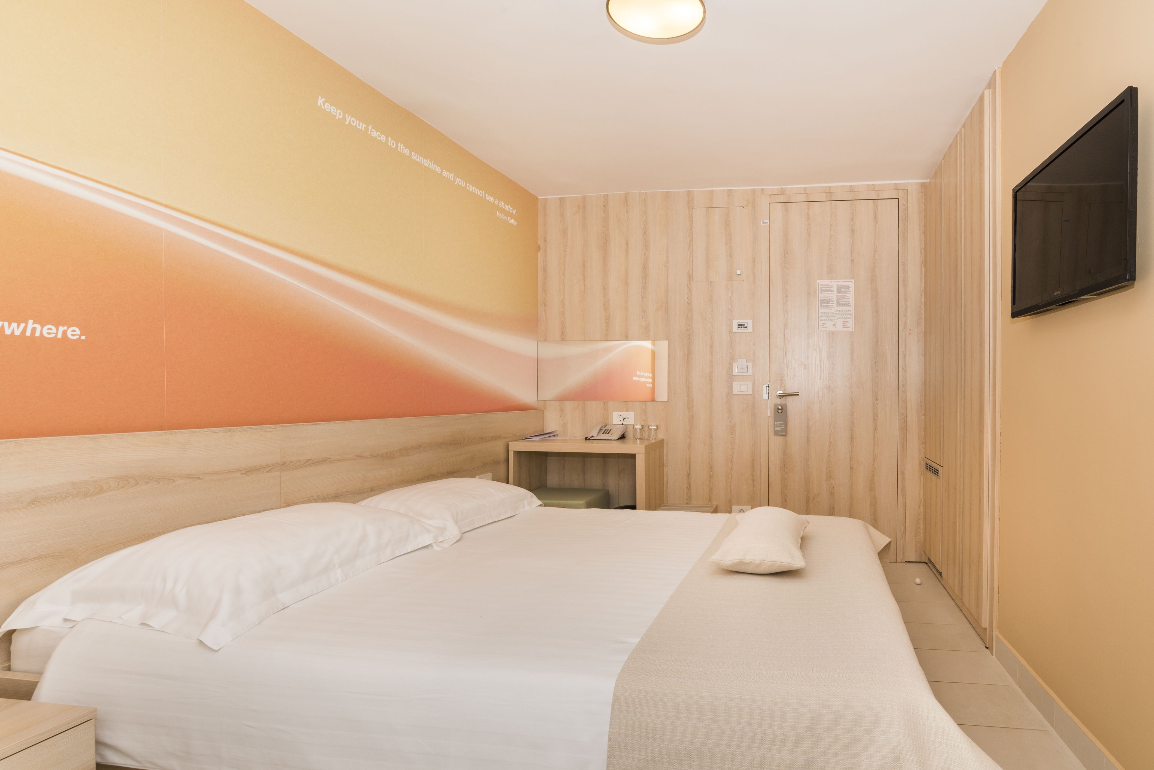 A hotel room with a large bed, a bedside table, a lamp, a TV mounted on the wall, and a wooden door.
