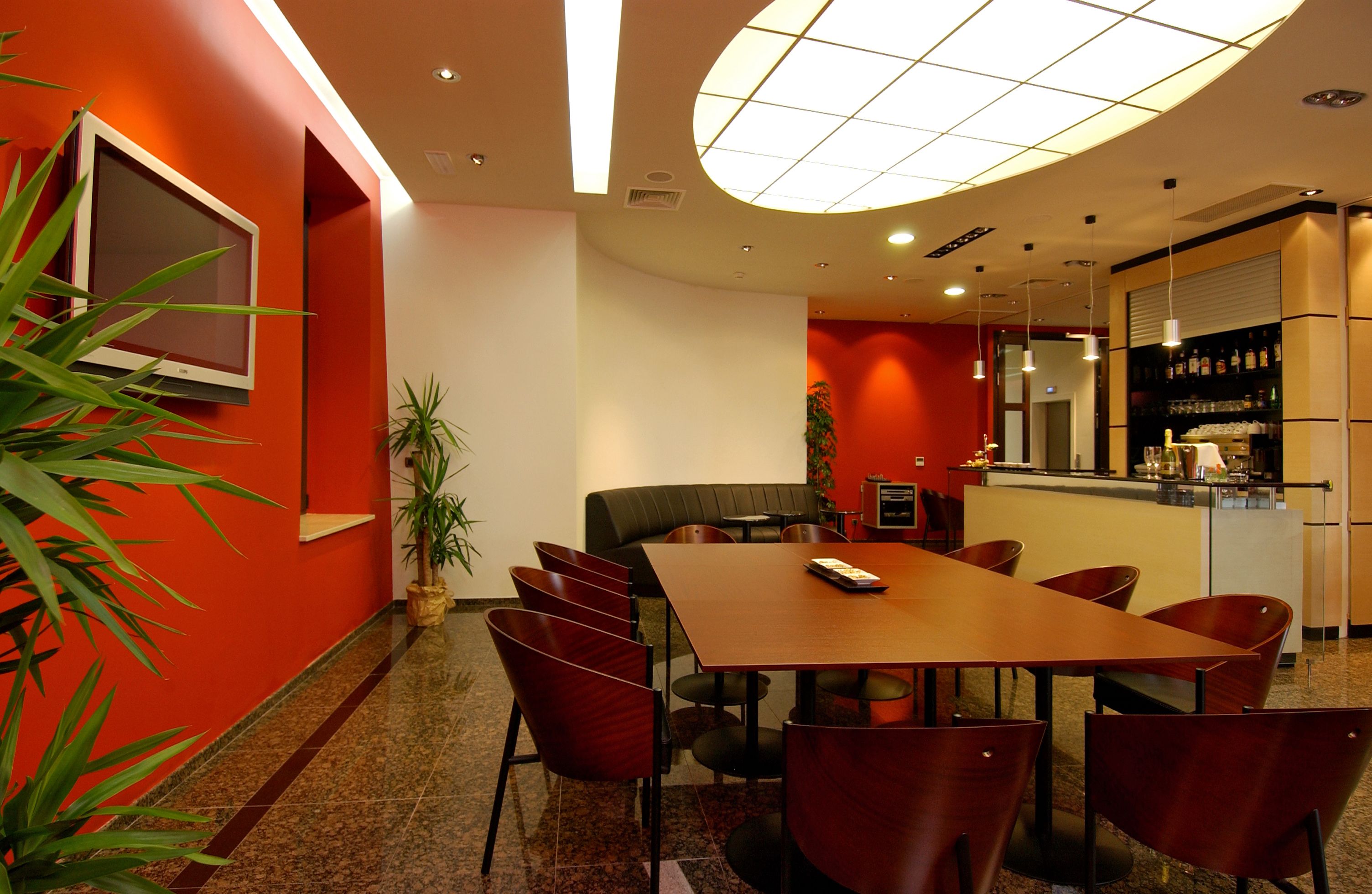 A modern conference room with a long table and red chairs, a small seating area, and a kitchen area with a counter and shelves.