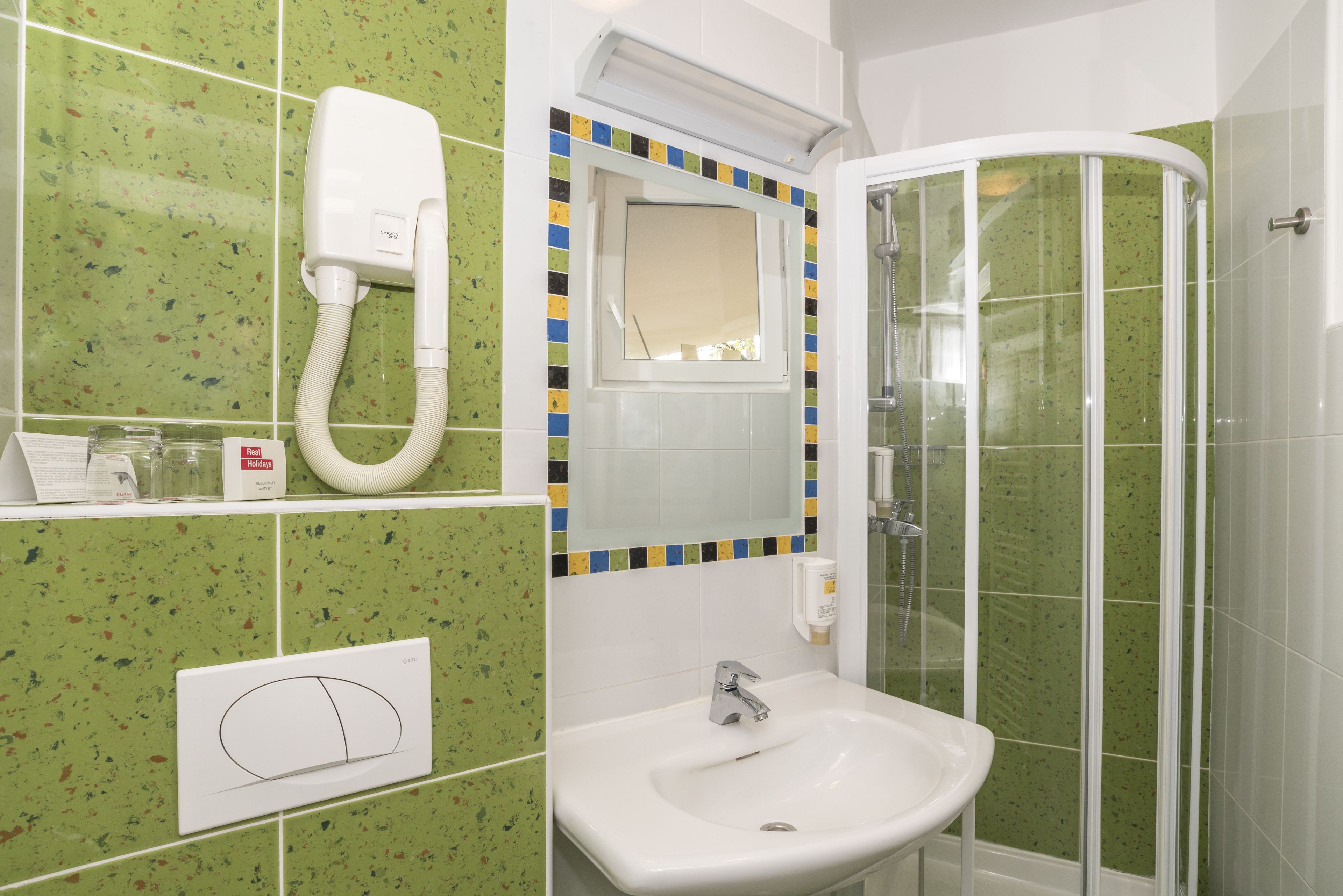 Een badkamer met groen gespikkelde tegels, een wit wastafelmeubel, een douche met glazen wanden en een spiegel met een kleurrijke omlijsting.