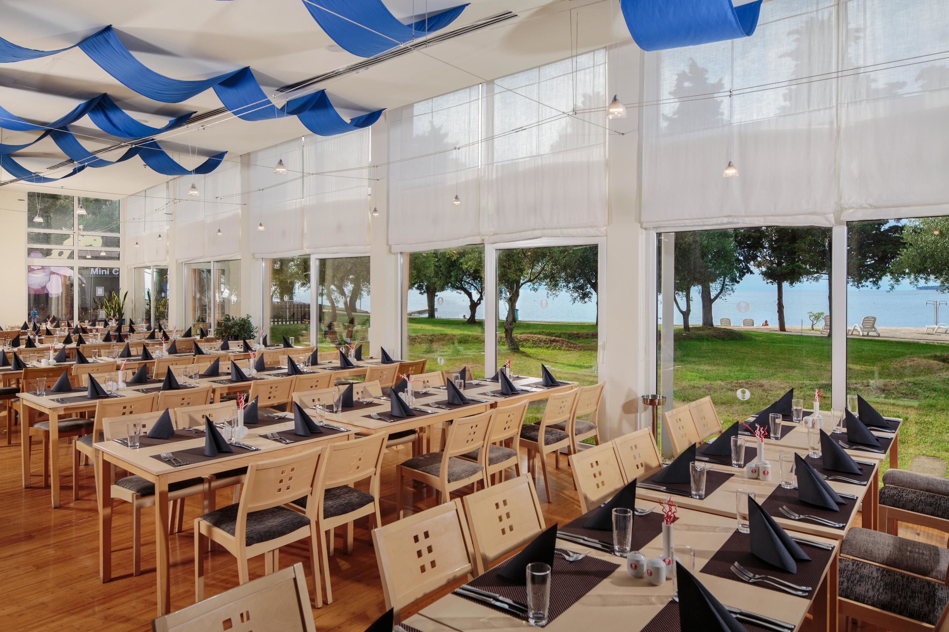 A restaurant with multiple tables set for dining, large windows offering a view of the sea, and a ceiling adorned with blue fabric.