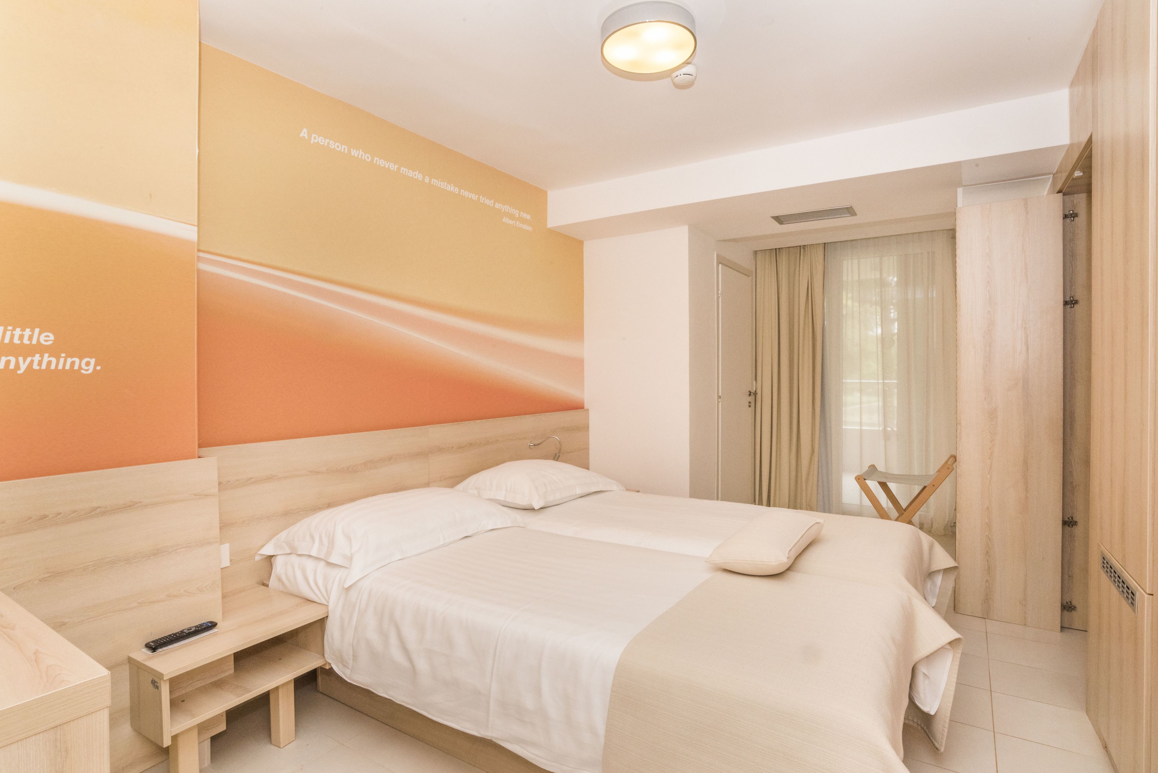 A modern hotel room with a wooden headboard, a bed with white linens, a bedside table with a remote control, and a wall with motivational text.