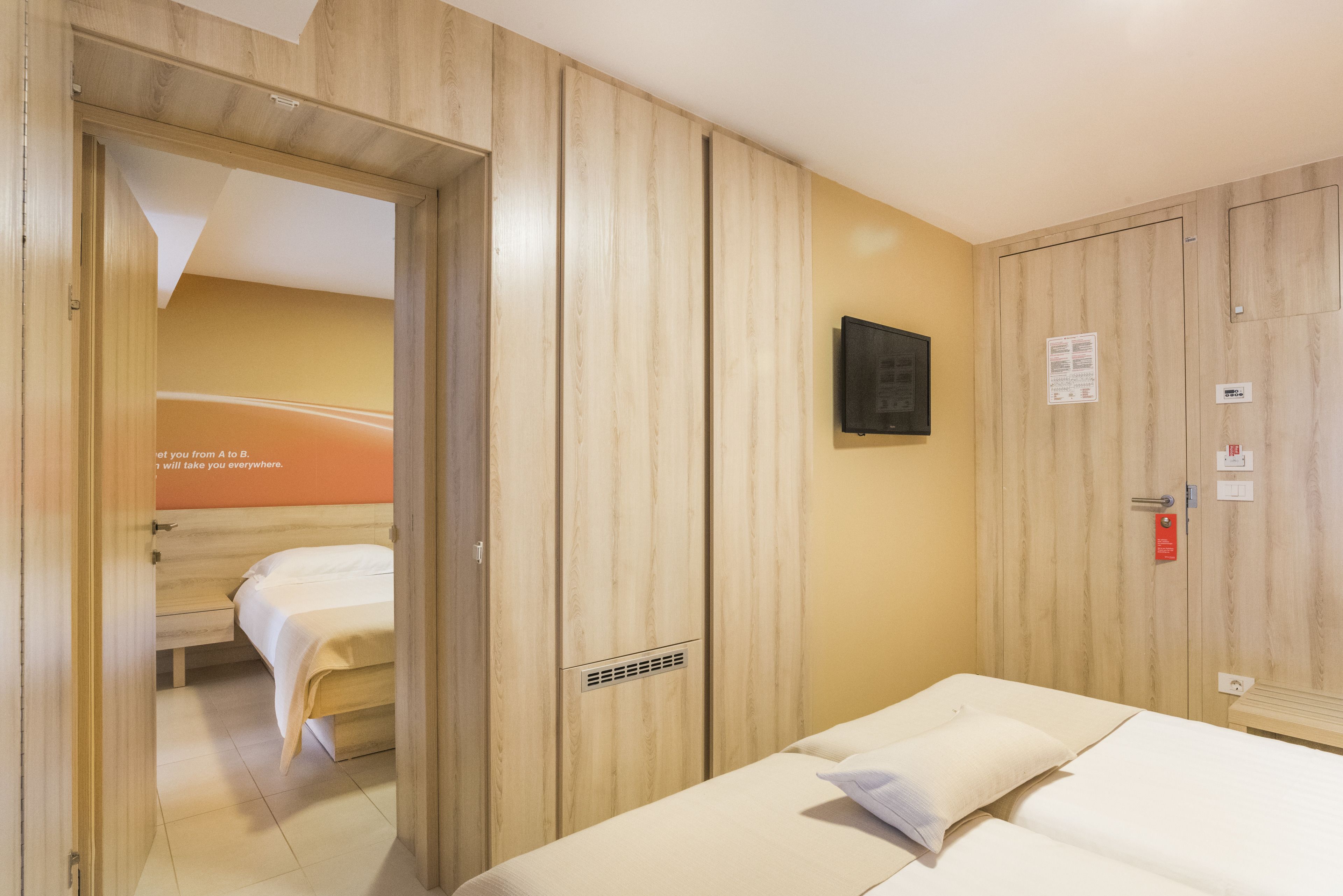 A hotel room with a wooden wardrobe, a bed with white bedding, a small bedside table, and a wall-mounted TV.