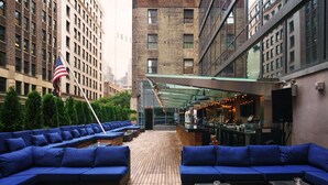 Un bar en la azotea con asientos azules, una bandera estadounidense y una zona de bar con iluminación ambiental.