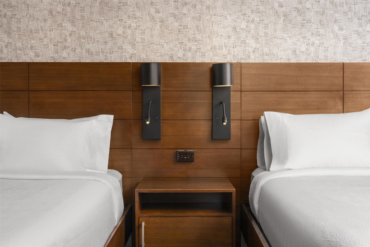 Habitación de hotel con dos camas, cabecero de madera, mesita de noche y dos lámparas de pared.